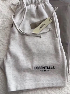 Grå Essentials Fear of God shorts - Snygga ljusgrå shorts från Essentials Fear of God med broderad logga på benet. De har elastisk midja med snörning och är tillverkade i mjuk bomull för maximal komfort. Perfekta för chill dagar eller streetwear-stil.