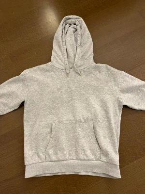 Ljusgrå hoodie med magficka - Säljer en enkel och stilren ljusgrå hoodie med huva och dragsnören. Hoodien har en stor magficka framtill och ribbade muddar vid ärmslut och nederkant. Perfekt för en avslappnad och bekväm stil.