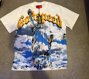 Vit t-shirt med frihetsgudinnan och tryck - Säljer en vit t-shirt med stort tryck av frihetsgudinnan i blått, svart och gult på framsidan samt texten 'Godspeed' i gul gotisk stil. Baksidan har en stor gul symbol och texten 'MANIFESTED IN MMXIII'. Klassisk passform och rund hals.