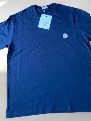Mörkblå t-shirt från Moncler - Snygg mörkblå t-shirt från Moncler med klassisk rund hals och diskret Moncler-logga på bröstet. Tillverkad i mjuk bomull som känns skön mot huden. Perfekt till jeans eller shorts för en clean och stilren look.