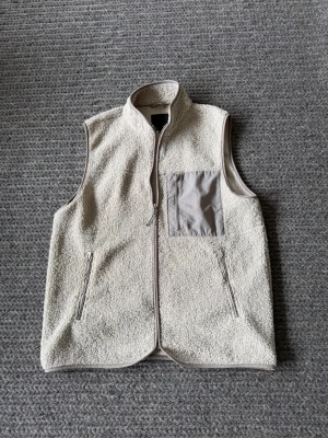 Beige fleeceväst med bröstficka - Säljer en beige fleeceväst med dragkedja framtill och två sidofickor med dragkedja. Västen har en kontrasterande bröstficka i grått tyg med dragkedja och hög krage. Perfekt för lager-på-lager och riktigt skön att ha på sig.