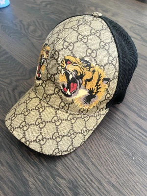 Gucci keps med tigertryck M - Snygg keps från Gucci med klassiskt GG-mönster i beige och brunt samt två tigerhuvuden framtill. Baksidan är i svart mesh för extra ventilation. Storlek M. Se etiketter som styrker äkthet!