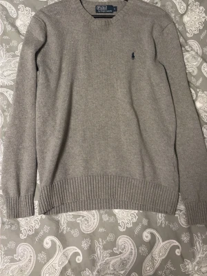Polo Ralph Lauren tröja - Klassisk grå stickad tröja från Polo Ralph Lauren med rund halsringning och diskret broderad logga på bröstet. Tröjan har ribbstickade muddar vid ärmslut och nederkant. Perfekt för lager-på-lager och enkel att matcha. Den är i fint skick utan defekter hör gärna av er vid fler frågor