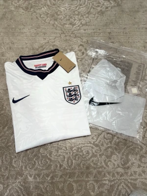 Vit England fotbollströja Nike - Säljer en vit England fotbollströja från Nike med broderat landslagsemblem och stjärna på bröstet. Tröjan har marinblå och röda detaljer i kragen samt Nike-logga. Tillverkad i Dri-FIT-material som andas. Perfekt för fotbollsträning eller match.