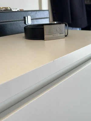 Svart skärp med silverdetalj från BOSS - Snyggt svart skärp från BOSS med en stilren, rektangulär silverspänne där BOSS-loggan är ingraverad. Skärpet har en klassisk look och passar perfekt till jeans eller kostymbyxor. Materialet är slätt skinn och spännet är i metall.