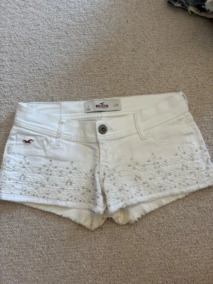 Vita broderade shorts från Hollister - Säljer ett par vita shorts från Hollister med broderade detaljer framtill och råa kanter. Shortsen har låg midja, klassiska fickor och knappstängning. Perfekta för sommaren och ger en fräsch och avslappnad look.💓