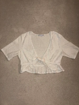 Vit bolero med knyt och volang - Söt vit bolero med knyt framtill och volangkant. Toppen har korta ärmar och är tillverkad i ett spetsliknande material med små hålmönster. Perfekt för en trendig och luftig look.
