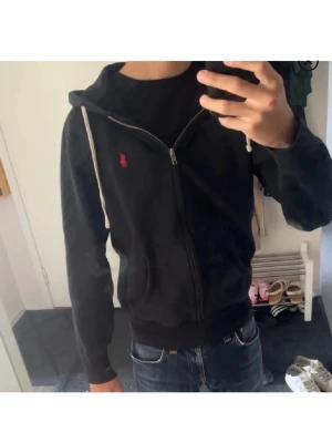 Svart zip hoodie från Polo Ralph Lauren - Säljer en svart hoodie med dragkedja från Polo Ralph Lauren. Hoodien har vit dragsko i huvan, två fickor framtill och den klassiska röda loggan broderad på bröstet. Perfekt till jeans och enkel att matcha med andra plagg.