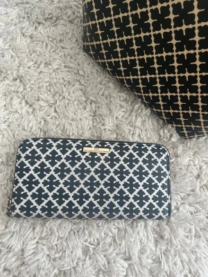 By Malene Birger liknande Svartvit mönstrad  - Snygg svartvit plånbok med grafiskt mönster och guldfärgad detalj framtill. Plånboken har dragkedja runtom och är tillverkad i ett tåligt material som liknar konstläder. Perfekt för dig som gillar stilrena accessoarer med en twist.