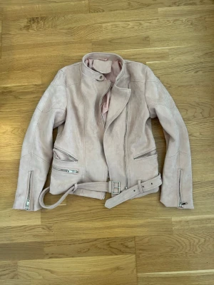 Ljusrosa mockajacka med bälte - Snygg ljusrosa mockajacka i bikerstil med silvriga dragkedjor och bälte i midjan. Jackan har ståkrage med tryckknapp och två snedställda fickor framtill. Perfekt för dig som vill ha en trendig och stilren look.