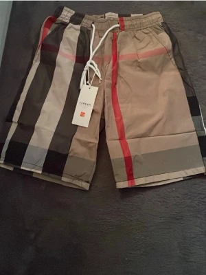 Burburry short - Snygga shorts med beige, svarta, gråa och röda rutor samt vit snörning i midjan. De har elastisk midja och ett klassiskt rutigt mönster som sticker ut. Perfekta för sommaren och ger en trendig vibe till din outfit.