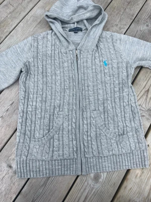 Grå kabelstickad zip hoodie - Snygg grå kabelstickad hoodie från Polo Ralph Lauren med dragkedja framtill och två fickor. Tröjan har ribbade muddar, huva och en blå broderad logga på bröstet. Perfekt för dig som gillar en avslappnad men stilren look.