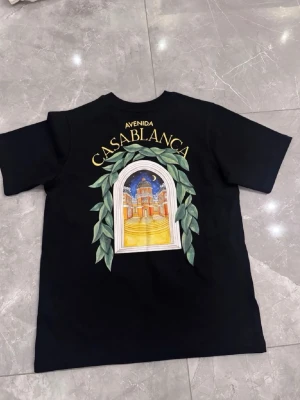 Svart t-shirt med tryck Casablanca - Säljer en svart t-shirt med färgglatt tryck av ett fönster och byggnad samt texten 'Avenida Casablanca' på både fram- och baksidan. T-shirten har korta ärmar och en klassisk passform. Perfekt för dig som gillar unika prints och streetwear vibes.