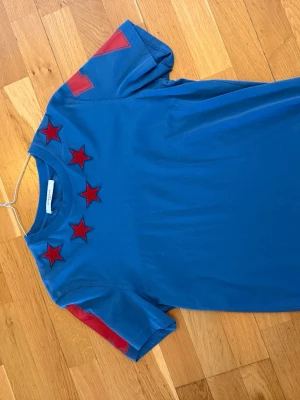 Blå Givenchy t-shirt med röda stjärnor - Säljer en blå t-shirt från Givenchy med röda stjärnor broderade runt halsen och röda detaljer på ärmarna. T-shirten har en klassisk passform och är tillverkad i mjuk bomull. Perfekt för dig som vill sticka ut med en unik designerpiece.