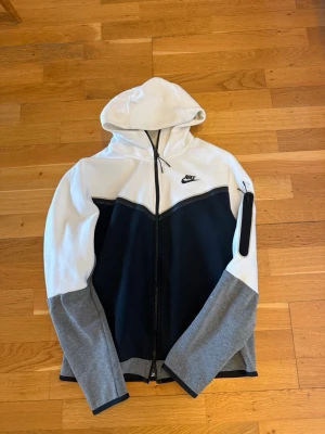 Nike hoodie med dragkedja och huva - Nike hoodie i vitt, svart och grått med dragkedja framtill och huva. Stora färgblock, svart Nike-logga på bröstet och en praktisk ficka med dragkedja på ena ärmen. Tillverkad i mjukt material, perfekt för en sportig och avslappnad stil.