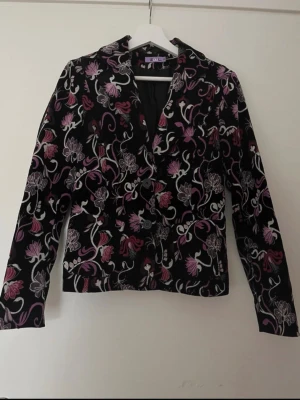 Blommig kavaj från Zoul - Snygg svart kavaj från Zoul med blommigt mönster i lila, rosa och vitt. Kavajen har klassisk krage, knäppning framtill. Perfekt för dig som vill sticka ut med en unik och färgglad stil.