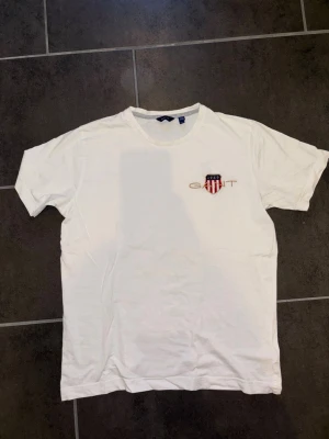 Vit t-shirt från Gant med broderad logga - Säljer en klassisk vit t-shirt från Gant med broderad logga och sköld på bröstet. T-shirten har rund halsringning och korta ärmar. Perfekt till jeans eller shorts för en clean och stilren look. Storlek S