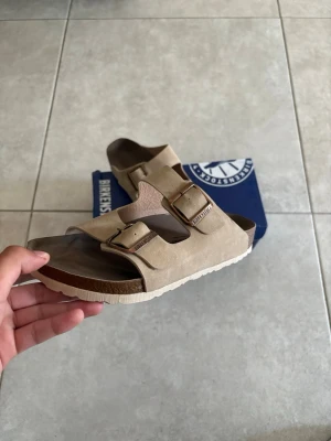 Beige Birkenstock sandaler (Arizona) - Säljer ett par klassiska beige sandaler från Birkenstock med två justerbara remmar och metallspännen. Ovandelen är i mockaliknande material och sulan är vit med korkdetaljer. Perfekta för en avslappnad och stilren look.