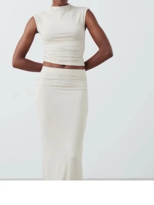 Beige bodyset med hög hals från ginatricot  - Snyggt och stilrent bodyset i beige med ärmlös topp och lång kjol. Toppen har hög hals och smickrande drapering i midjan. Setet är figurnära och tillverkat i ett mjukt, stretchigt material som sitter bekvämt mot kroppen.