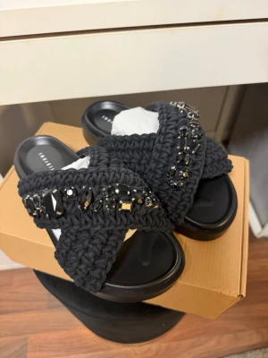 Svarta sandaler med pärlor från Inuikii - Säljer ett par svarta sandaler från Inuikii med breda, stickade band i grovt garn och dekorativa svarta och silvriga pärlor. Sandalerna har en chunky sula och öppen tå, perfekt för dig som vill ha något unikt och trendigt till sommaren.