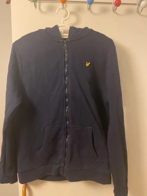 Mörkblå hoodie med dragkedja - Mörkblå hoodie med hel dragkedja framtill och två fickor. Har en huva och ett litet gult broderat märke på bröstet. Perfekt för en avslappnad och stilren look. Mjuk bomullskänsla och lång ärm.