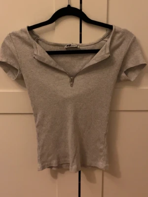 Ljusgrå ribbad topp med dragkedja - Säljer en ljusgrå ribbad topp med korta ärmar och en snygg dragkedja framtill som kan justeras för olika looks. Toppen har en tight passform. Perfekt till jeans eller kjol för en trendig vardagsstil.
