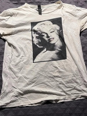 Vit t-shirt med Marilyn Monroe-tryck - Säljer en vit t-shirt med ett stort svartvitt tryck av Marilyn Monroe på framsidan. Klassisk passform och rund halsringning. Perfekt för dig som gillar ikoniska motiv och vill ha en stilren men ändå uttrycksfull look.
