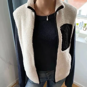 Vit teddyväst med svart ficka - Säljer en krämvit teddyväst med dragkedja och hög krage. Västen har en svart bröstficka med dragkedja som detalj och är gjord i mjukt, fluffigt material. Perfekt för lager på lager och ger en cool, avslappnad look. Storlek s💓