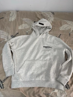 Ljusgrå Essentials Fear of God hoodie - Snygg ljusgrå hoodie från Essentials Fear of God med svart logga på bröstet och stor text på ryggen. Klassisk huva och magficka framtill. Mjuk bomullsblandning och ribbade muddar vid ärmslut och nederkant. Perfekt för en avslappnad streetwear-look.