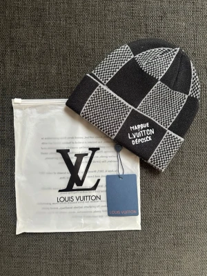Svartvit rutmönstrad mössa - Snygg svartvit mössa med rutigt mönster och texten broderad framtill. Mössan är stickad och har en uppvikt kant. Perfekt accessoar för dig som vill ha en exklusiv och trendig look. Aldrig använd.