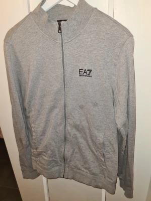 Grå zip-hoodie från EA7 Emporio Armani - Snygg grå hoodie med dragkedja från EA7 Emporio Armani. Tröjan har en stilren design med logga på bröstet, ribbade muddar och två fickor framtill. Perfekt för en avslappnad och trendig look. Tröjan har 2 fläckar på sig som borde gå bort med någon fläckborttagning. Kan behövas en strykning. 