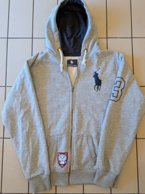 Grå zip hoodie från Polo Ralph Lauren - Snygg grå hoodie med dragkedja från Polo Ralph Lauren. Med en stor polologga på bröstet. "3"an på vänster arm har tappat lite tråd på toppen (Kolla sista bilden) men problemet är fixat och syns knappt längre