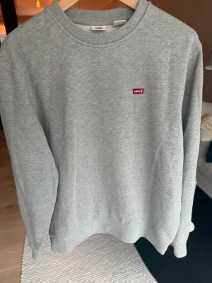 Grå sweatshirt från Levi's - Klassisk grå sweatshirt från Levi's med rund halsringning och diskret röd logotyp på bröstet. Tröjan har ribbade muddar vid ärmslut och nederkant, perfekt för en avslappnad och stilren look.