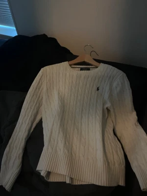 Vit kabelstickad tröja Polo Ralph Lauren - Kabelstickad vit tröja från Polo Ralph Lauren med rund halsringning och diskret logga broderad på bröstet. Långa ärmar och breda ribbade muddar vid ärmslut och nederkant. Klassisk och tidlös design som passar till allt.