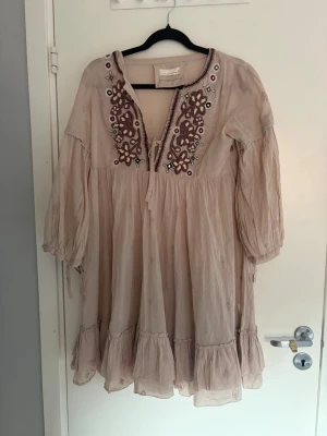 Beige broderad klänning Odd Molly - Supersöt beige kortklänning från Odd Molly med broderade detaljer i vinrött och vitt längs ringningen. Klänningen har v-ringad hals, trekvartsärm och volangkant nedtill. Perfekt för sommarens alla tillfällen!