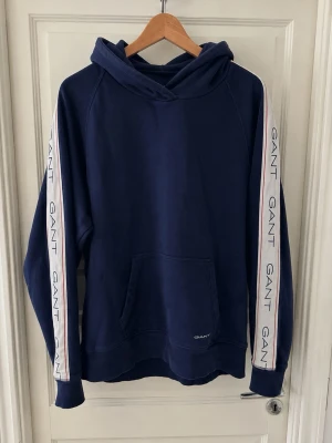 Marinblå hoodie från GANT - Snygg marinblå hoodie från GANT med huva och känguruficka. Ärmarna har vita band med GANT-logga och tunna röda linjer längs sidorna. Tillverkad i mjukt bomullsmaterial, perfekt för en avslappnad och sportig stil.