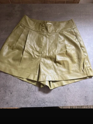 Gröna shorts EDITED 36 - Snygga gröna shorts i skinnimitation med två knappar framtill och veckade detaljer. Shortsen har en stilren look och passar perfekt till en trendig outfit. I storlek 36
