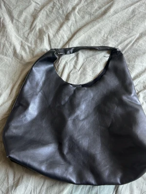 Svart slouchy axelväska i fejkläder - Stilren svart handväska i mjukt fejkläder med slouchy form och bred axelrem. Väskan har minimalistisk design utan synliga detaljer eller logga, perfekt för dig som gillar en clean look. Rymmer allt du behöver till vardagen och är enkel att matcha med olika outfits.