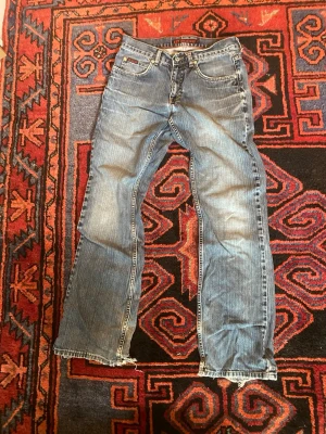 Blå raka jeans med slitna benslut - Ett par klassiska blå jeans med rak passform och slitna, fransiga benslut. Jeansen har fem fickor och tydliga sömmar. Perfekta för en avslappnad och trendig look. Materialet är denim i bomull.