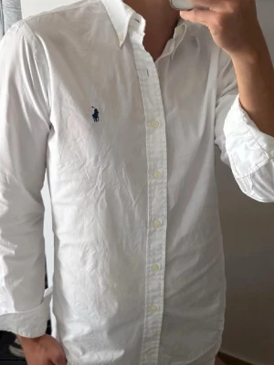 Vit skjorta med broderad logga - Klassisk vit skjorta med lång ärm och knappar framtill. Skjortan har en broderad marinblå logga på bröstet och en stilren krage. Tillverkad i bomull och passar perfekt till en clean och tidlös stil.