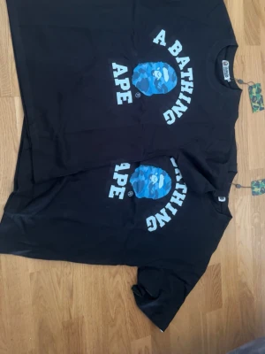 Svart BAPE t-shirt med blått camomönster - Säljer en svart t-shirt från A Bathing Ape med det klassiska BAPE-huvudet i blått camomönster och vit text. T-shirten har rund hals och korta ärmar, perfekt för en streetwear-look.
