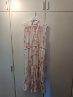 Desmond & Dempsey för H&M.Vit långklänning med palmmönster stl S - Långklänning från H&M med v-ringning och knappar framtill. Klänningen är vit med ljusrosa palmmönster, ärmlös och har volangdetaljer. Perfekt för sommarens varma dagar och har en avslappnad passform.