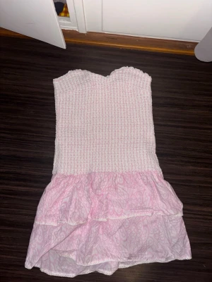 Rosa axelbandslös kortklänning Gina Tricot - Söt axelbandslös kortklänning från Gina Tricot i ljusrosa och vitt. Klänningen har smock upptill och volangig kjol med diskret mönster. Har aldrig använt