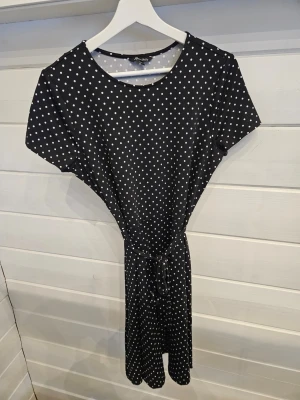 Svart prickig kortklänning med knyt - Svart kortklänning med vita prickar och knyt i midjan. Klänningen har rund halsringning och korta ärmar. Tillverkad i mjuk polyester med lite stretch för skön passform. Perfekt för dig som gillar en lekfull och stilren look.