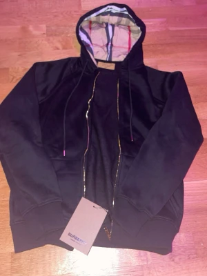 Svart hoodie från Burberry med huva - Snygg svart hoodie från Burberry med dragkedja och klassiskt rutigt foder i huvan. Hoodien har ribbade muddar, fickor framtill och justerbara snören i huvan. Perfekt för dig som vill ha en stilren och exklusiv look.