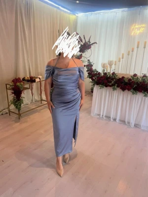 Ljusblå offshoulder långklänning - Elegant ljusblå långklänning med offshoulder och draperad design. Klänningen har en slits framtill och smickrande passform som framhäver figuren. Perfekt för speciella tillfällen och har en mjuk, glansig yta.