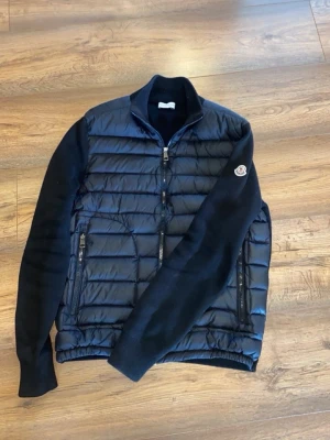 Svart cardigan från Moncler - Snygg svart pufferjacka från Moncler med quiltad front och rygg i glansigt material samt stickade ärmar. Jackan har två dragkedjefickor fram och en hög krage. Moncler-logga på ärmen ger en exklusiv touch. Perfekt för kalla dagar.