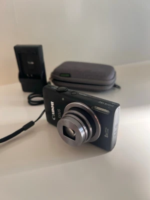 Canon IXUS 132 kompaktkamera svart - Snygg och smidig svart Canon IXUS digitalkamera med 8x optisk zoom och silverfärgat objektiv. Kameran har en klassisk rektangulär form och levereras med laddare, handledsrem och en grå skyddsväska. Perfekt för dig som vill ha en fin digitalkamera till sommaren📸☀️ två sista bilderna visar på 2 små repor