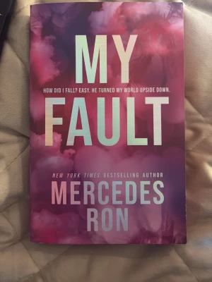 My Fault av Mercedes Ron - Säljer boken 'My Fault' av Mercedes Ron, en ungdomsroman på engelska. Omslaget har en rosa och lila färgskala med stora vita bokstäver. Boken handlar om Noah som flyttar till LA och möter sin nya bror Nicholas, vilket leder till drama, kärlek och spänning.