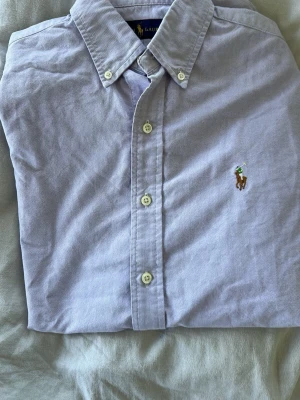 Ljuslila skjorta från Ralph Lauren XS - Klassisk ljuslila skjorta från Ralph Lauren med button-down krage och broderad logga på bröstet. Skjortan har långa ärmar och vita knappar framtill. Tillverkad i mjuk bomull som känns skön mot huden.